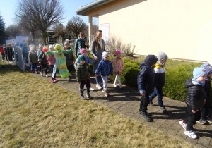 Dzieci wraz z paniami maszerują z wiosennymi plakatami, panią Wiosną pod budynkiem przedszkola.